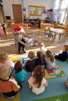 Zdjęcie przedstawia nauczyciela w czapce Mikołaja pokazującego dzieciom ilustracje z książki.
