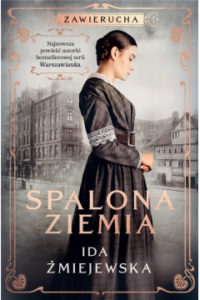Zdjęcie okładki książki pt. "Spalona ziemia" autorstwa Idy Żmijewskiej. Na pierwszym planie widoczna postać kobiety w długiej ciemnej sukni, w tle widać szare zabudowania.