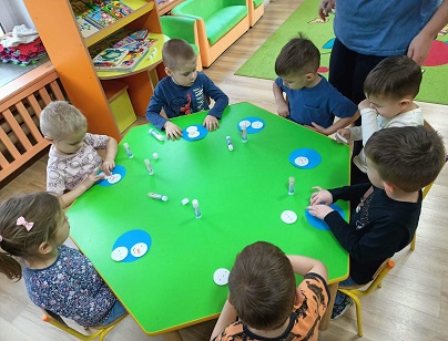 Zdjęcie przedstawia dzieci siedzące przy zielonym stoliku i wykonyjące prace plastyczne.