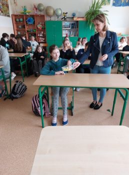 Na zdjęciu uczniowie siedzą w ławkach, kobieta stoi przy ławce i daje uczennicy pudełko z papierowymi kwiatami do wyboru.