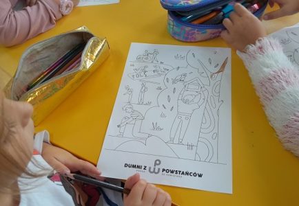 Na zdjęciu widoczna kartka z rysunkiem do kolorowania, leżąca na stoliku przy którym siedzą dzieci.