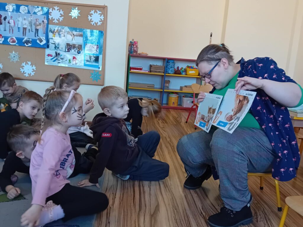 Na zdjęciu kobieta siedzi na małym krzesełku. Przed nią na dywanie siedzą dzieci i patrzą na obrazki, które kobieta pokazuje.