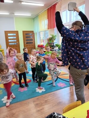 Na zdjęciu widać kobietę stojąca na jednej nodze, pokazuje ćwiczenia dzieciom.