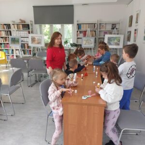 Na zdjęciu widać dzieci w sali przy biurkach, przed nimi papierowe elementy, kleje. Z lewej strony stoi kobieta w czerwonym swetrze. W głębi sali obrazy na sztalugach i regały z książkami.