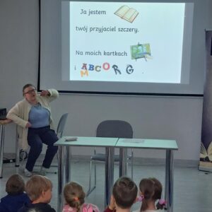 Na zdjęciu widoczna kobieta siedząca na krześle, za nią na ekranie wyświetlona plansza z wierszem i rysunkami, przed dwa złączone ze sobą stoliki oraz siedzące dzieci.