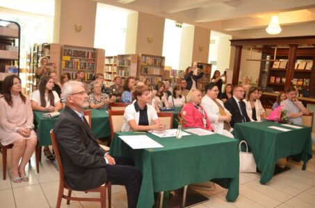 Zdjęcie przedstawia siedzących ludzi w sali, czy stolikach. Z tyłu widoczne regały z książkami.