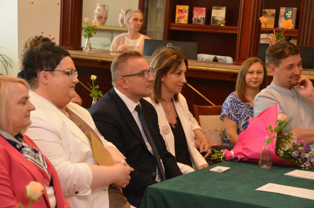 Na zdjęciu przy stoliku widać siedzące osoby. Na stoliku leży bukiet kwiatów.