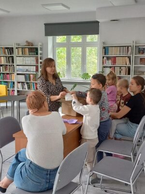 Na zdjęciu widać dzieci przy stoliku. Na pierwszym planie kobieta odwrócona tyłem, Druga stoi przy stoliku, przed dziećmi i coś wyjmuje z paczki. W tle białe regały z książkami.