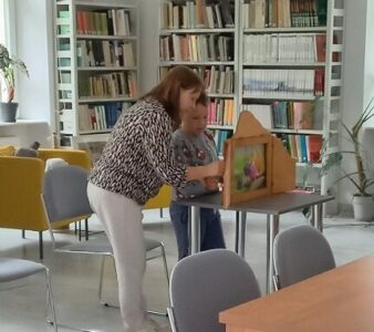 Na zdjęciu widać kobietę i chłopca stojących przy stoliku, na którym stoi drewniana skrzyneczka z ilustracjami w środku. Kobieta coś pokazuje chłopcu.