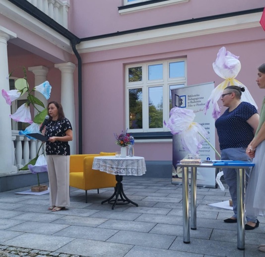 Na zdjęciu widać kobiety stojące przed budynkiem. w głębi widać duże papierowe kwiaty, żółty fotel, stolik z bukietem. Po prawej stronie drugi stolik.