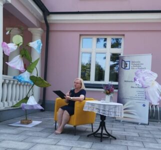 Na zdjęciu kobieta siedzi w fotelu i czyta. Obok stolik przykryty obrusem i bukiem. Z tyłu baner instytucji i ściana różowego budynku z oknem.
