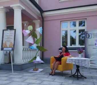 Na zdjęciu widać kobietę siedzącą w żółtym fotelu i czytającą, w tle ściana budynku, obok duży kwiat robiony, z tyłu baner instytucji.