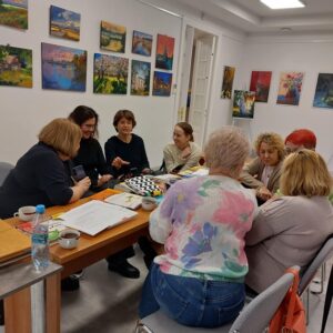 Na zdjęciu widać grupę kobiet siedzących wokół stołu , nachylonych ku sobie, przed nimi na stole rozłożone książki.