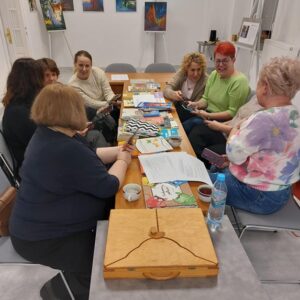 Na zdjęciu widoczna grupa kobiet siedzących w sali przy długim stole. Przed nimi rozłożone książki.