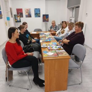 Na zdjęciu widać grupę ludzi w sali, siedzących przy stole, na którym leżą książki, w tle widać wiszące na ścianie obrazy.