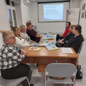 Na zdjęciu osoby siedzące przy stole na którym rozłożono książki, w tle ekran projektora.