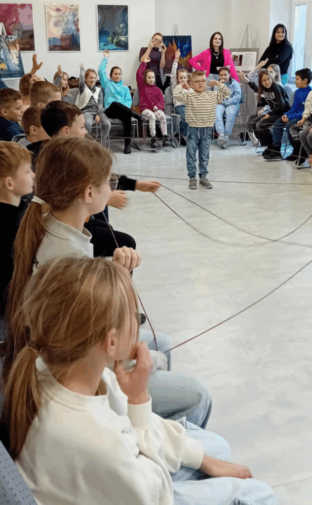 Na zdjęciu widać dzieci siedzące po bokach sali, pomiędzy nimi rozciągnięta nić w formie pajęczyny.