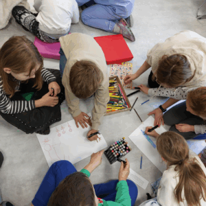 Zdjęcie przedstawia dzieci rysujące kredkami na kartkach na podłodze.