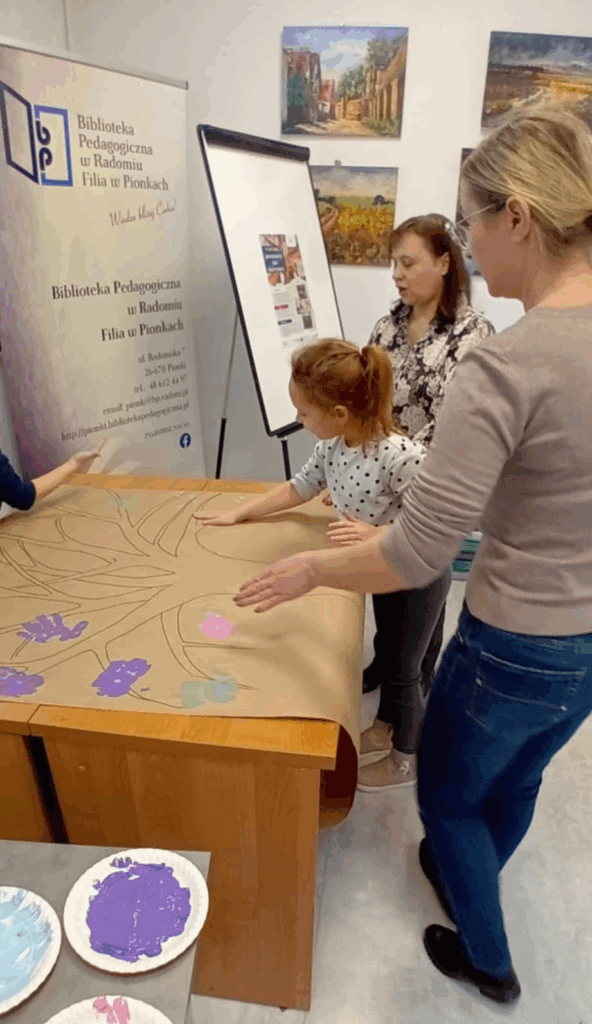 Na zdjęciu widać kobietę stojącą przy stoliku z rozłożonym kartonem, na którym narysowane jest drzewo, dziewczyna ma położoną rękę na kartonie.