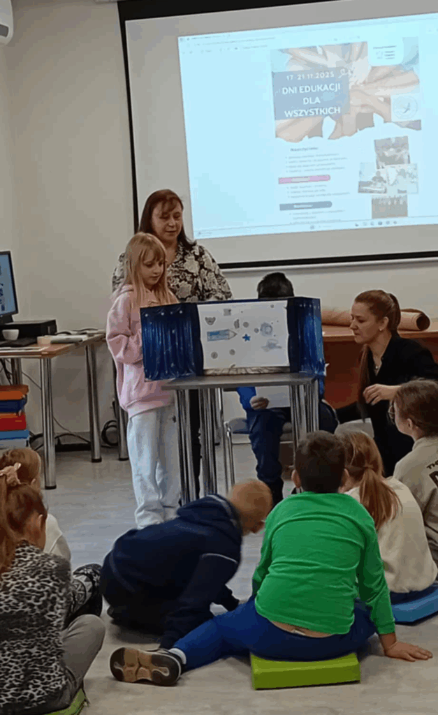 Na zdjęciu widać dzieci siedzące na podłodze, przed nimi na stoliku rozłożony teatrzyk papierowy z włożonymi rysunkami, obok stoi dziewczynka, kobieta. Z tyłu widać głowę chłopce i drugą kobietę.