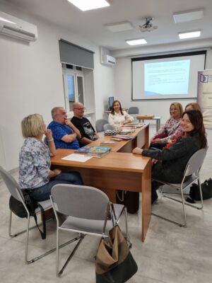 Na zdjęciu grupa osób siedzących przy stole w sali konferencyjnej. Na stole ułożone książki, w tle ekran projektora.
