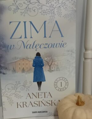 Zdjęcie okładki książki "Zima w Nałęczowie". W scenerii zimowej na pierwszym planie kobieta stojąca tyłem w niebieskim płaszczu, w tle beżowe domy. U góry tytuł książki.