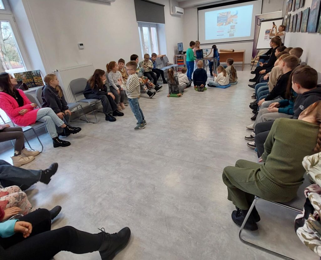 Na zdjęciu widać grupę dzieci siedzących na krzesłach, po bokach sali, przed nimi przy małym stoliku stoją uczniowie, część siedzi na podłodze. Na stoliku ustawiony karton z rysunkami.