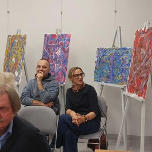 Na zdjęciu widać ludzi siedzących w sali wystawowej, z tyłu stoją sztalugi z obrazami