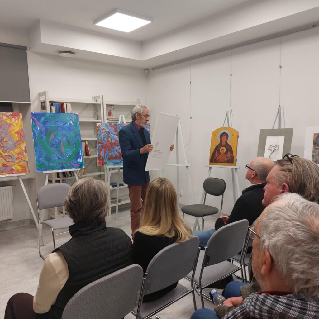 Na zdjęciu sala wystawowa. W centralnej części mężczyzna trzyma obraz, przed nim siedzą ludzie.