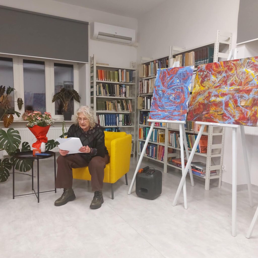 Na zdjęciu widać siedzącą kobietę w fotelu, w rękach trzyma kartki i czyta, za nią w głębi regały z książkami.