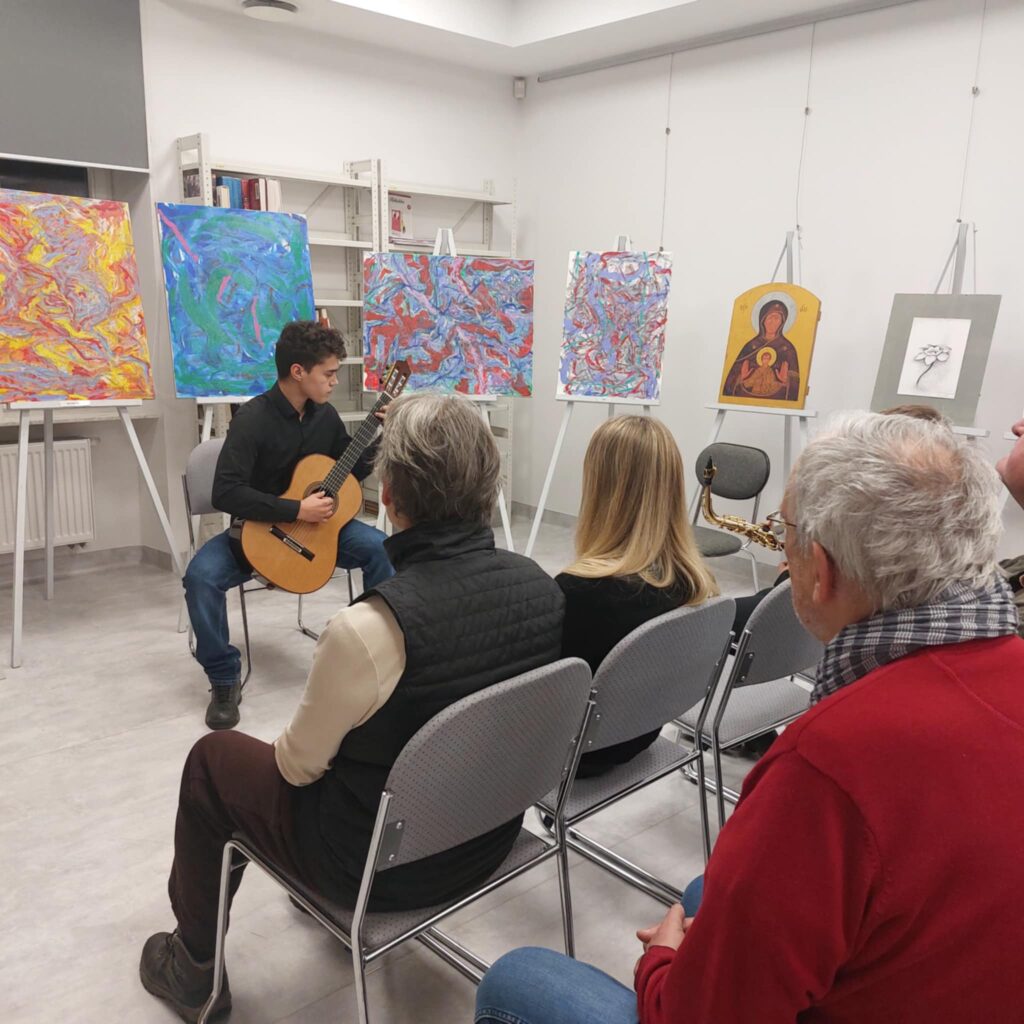 Na zdjęciu widać chłopca grającego na gitarze, za nim ustawione obrazy na sztalugach, przed nim siedzący ludzie i słuchający.