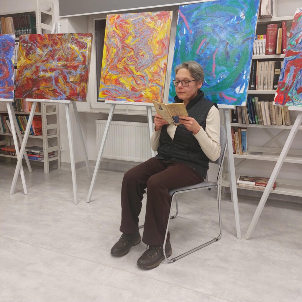 Na zdjęciu w centralnej części kobieta siedząca kobieta z książką w rękach, za nią stoją sztalugi z kolorowymi obrazami.