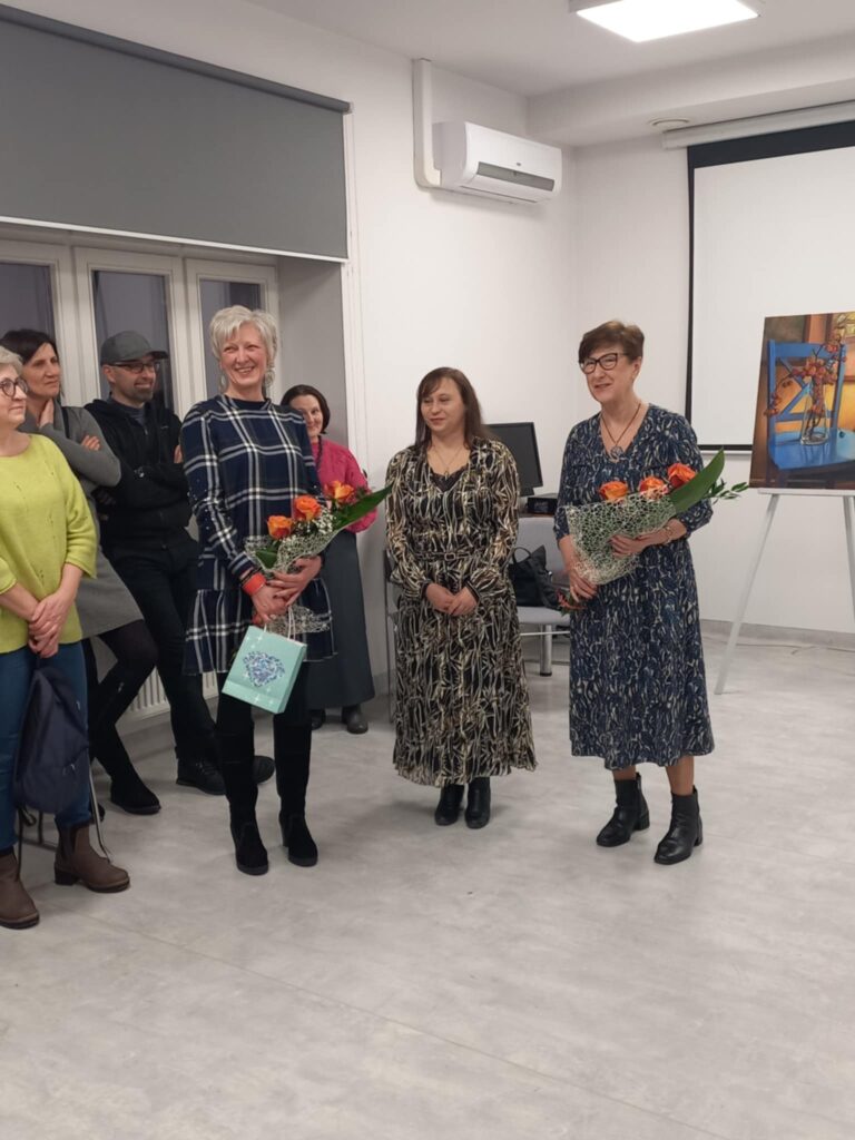 Na zdjęciu widoczne osoby stojące w grupie w sali, dwie trzymają w rękach bukiety kwiatów.