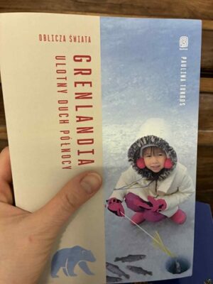 Zdjęcie okładki książki "Grenlandia. Ulotny duch północy". Po prawej stronie widać dziecko wyciągające rybę z przerębli. Obok na lodzie leżą inne ryby. Po lewej stronie na beżowym tle u góry tytuł książki, na dole mały niedźwiedź,