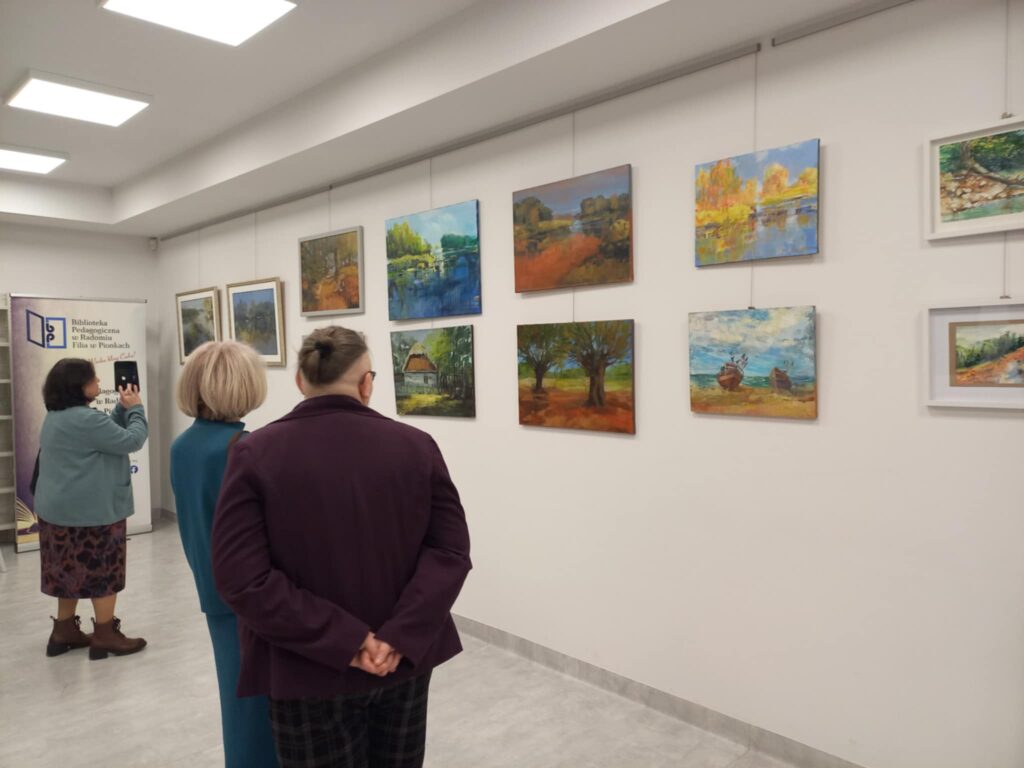 Na zdjęciu widoczne osoby oglądające obrazy wiszące na ścianie, w głębi widoczny baner instytucji.
