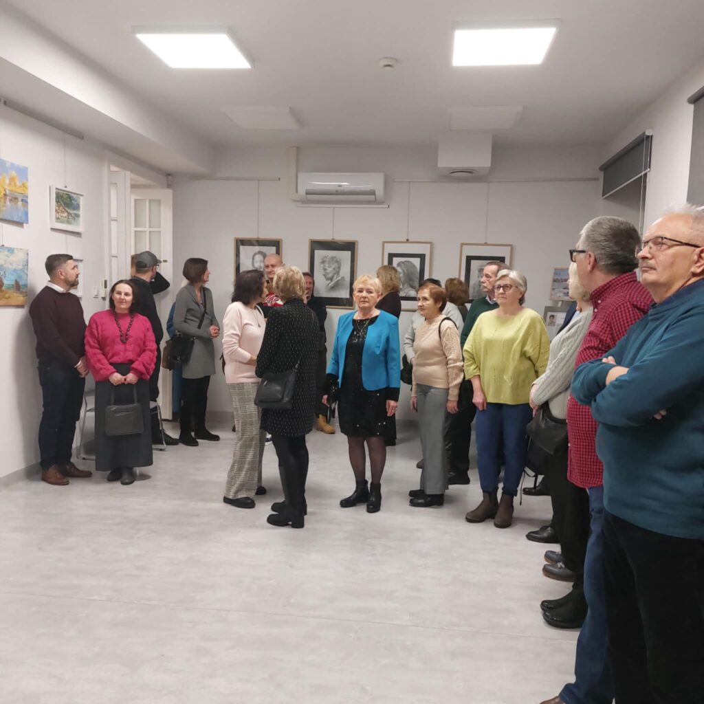 Na zdjęciu widoczne osoby zgromadzone w sali, na ścianach wiszą obrazy.