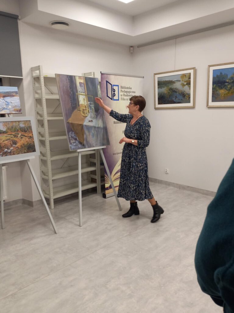 Na zdjęciu widoczna kobieta pokazująca obraz stojący na sztaludze, z nią na ścianie wiszą obrazy.