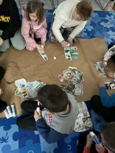 Na zdjęciu dzieci w sali przyklejają kolorowe obrazki na duży arkusz papieru z narysowanymi konturami.