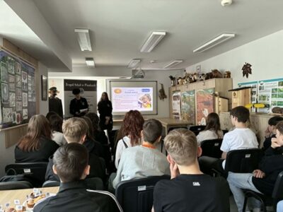 Na zdjęciu widać młodzież siedzącą w sali. Przed nimi przy ekranie stoją dwaj chłopcy i dziewczyna.