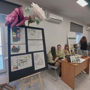 Na zdjęciu fragment sali, po lewo stoi tablica z planszami, na niej papierowe duże kwiaty, po prawo przy biurku siedzą 3 dziewczyny. Za nimi plansza informacyjna.