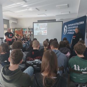 Na zdjęciu widoczna młodzież oglądająca prezentację, po bokach widocznie prowadzący spotkanie.