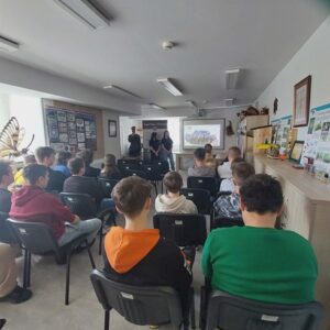 Zdjęcie przedstawia salę, a w niej młodzież słuchającą prezentacji wyświetlanej na ekranie przez prowadzących uczniów.
