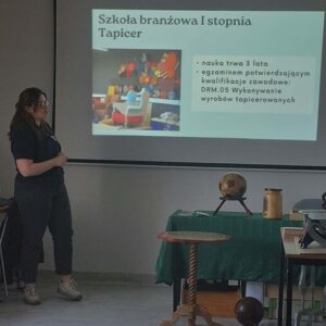 Na zdjęciu widać kobietę patrzącą na ekran z wyświetloną informacją. po prawej stronie widać stół przykryty zielonym materiałem, na nim ustawione różne przedmioty z drewna. Przed nim stoi mały drewniany stolik na 1 nóżce.