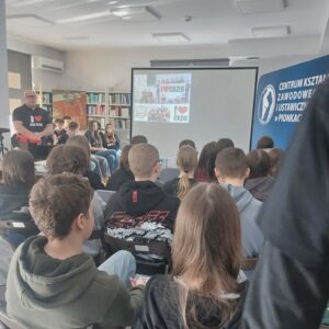 Na zdjęciu widać siedzącą młodzież w sali, przed nimi ekran z pokazywaną prezentacją i prowadzący.
