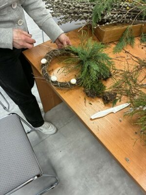 Na zdjęciu widać stolik z różnymi gałązkami, przy nim stoi osoba i robi wieniec.