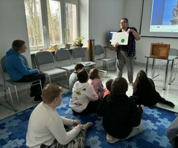 Na zdjęciu fragment sali. Na dywanie siedzą dzieci, przed nimi kobieta pokazuje plansze.