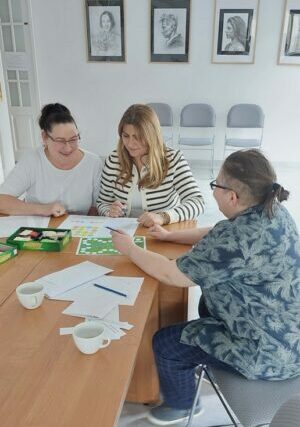 Na zdjęciu widoczne trzy siedzące kobiety przy biurku, przed nimi rozłożone kartki.